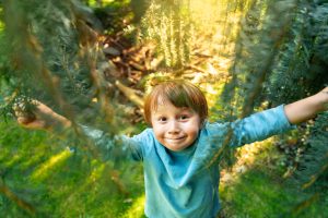 Portrait of a happy little boy playing in hide and seek in a park,  child pushing the branches of a tree and smiling, looking at the camera. Closeness to nature.